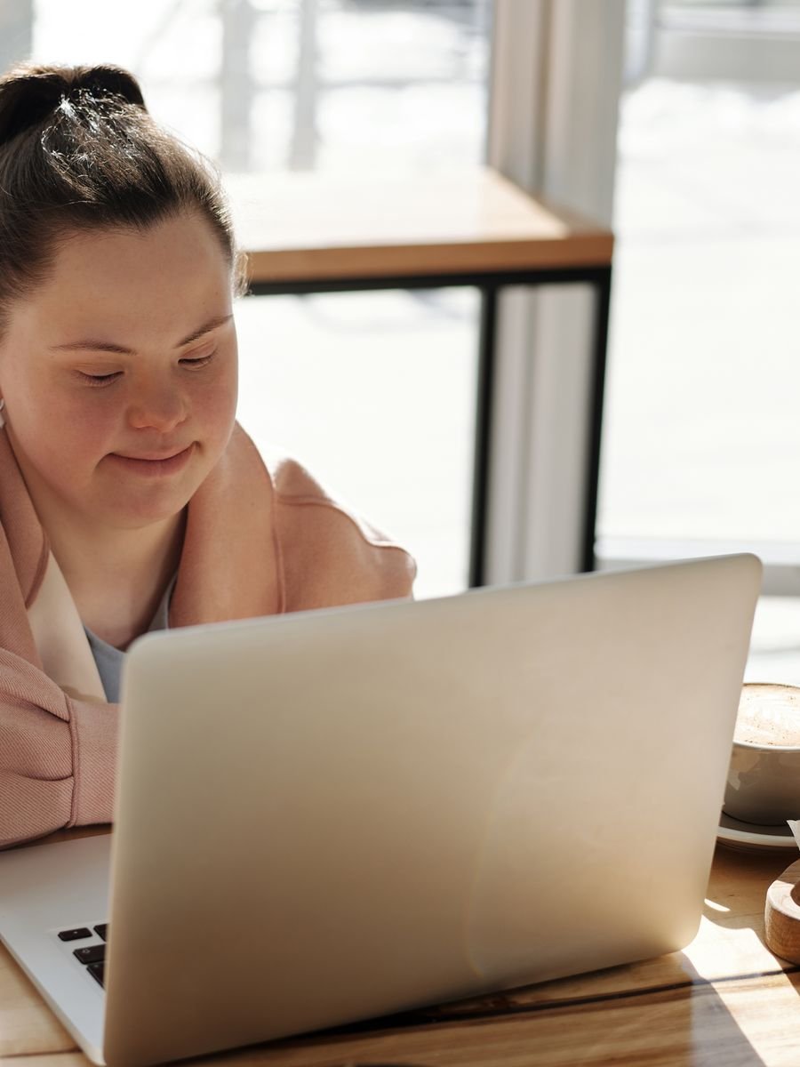 A young woman with Down syndrome working on a laptop in a sunlit cafe