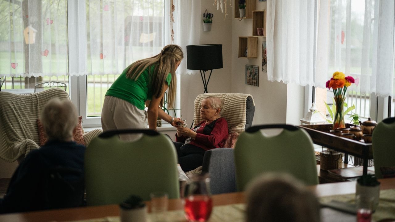 A caregiver sitting with an older woman in her armchair by the window